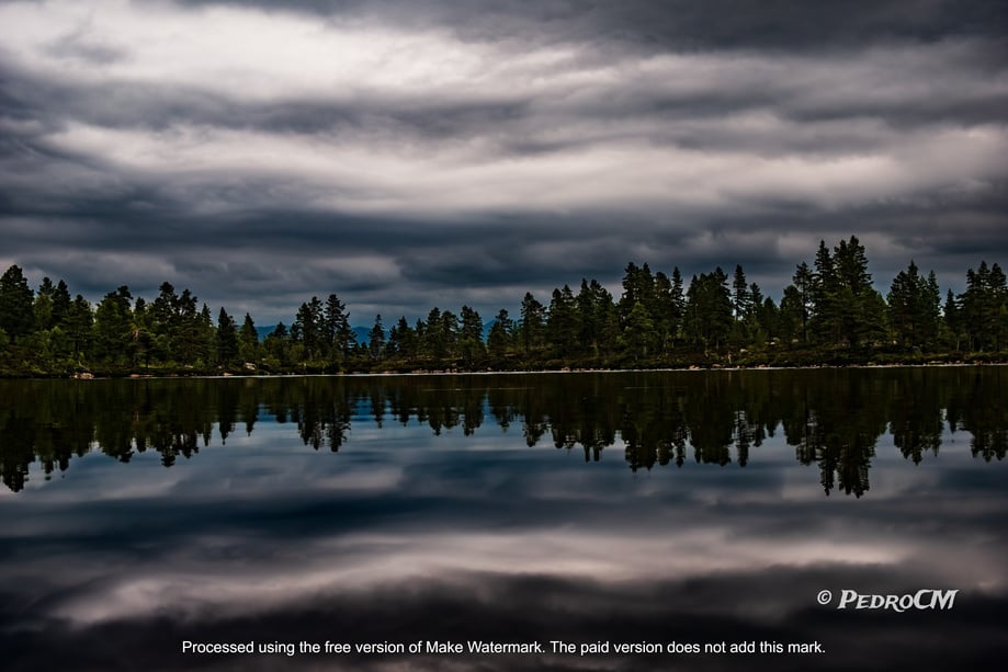 a dark stormy sky is reflected in the water of a lake with a treeline